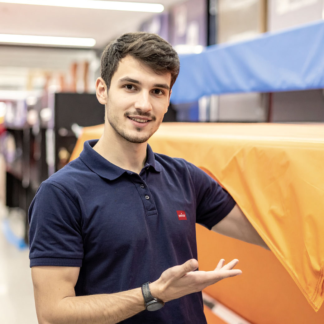 Junger Mann mit dunklem T-Shirt, lächelnd und gestikulierend vor einem bunten Hintergrund.