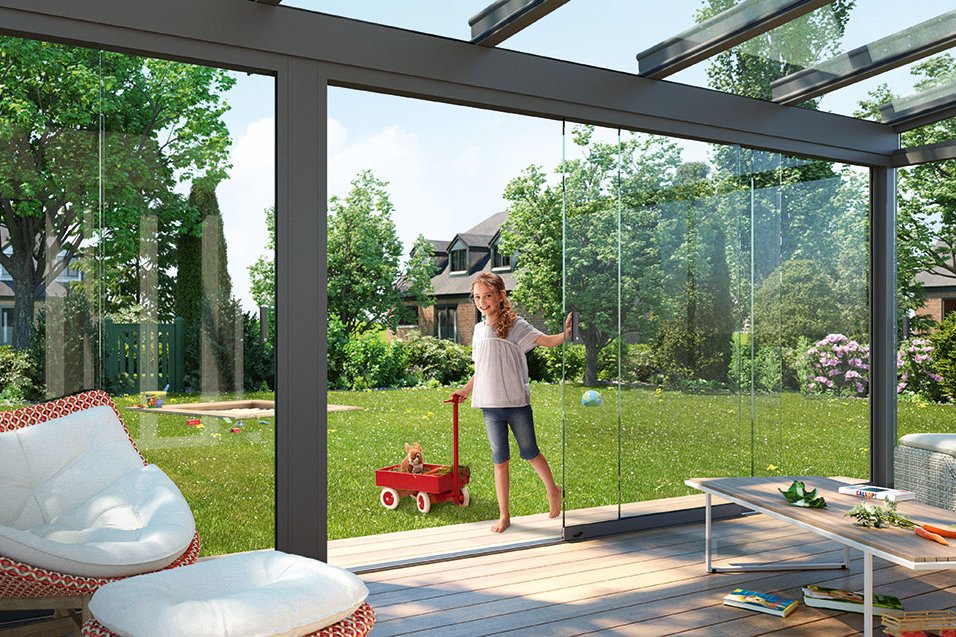 Sonniger Wintergarten mit Glas-Schiebewänden, Blick auf eine grüne Gartenlandschaft. Sonniger Wintergarten mit Glas-Schiebewänden, Blick auf eine grüne Gartenlandschaft.