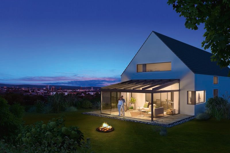 Ein modernes Haus mit Glas-Schiebewänden, umgeben von Grün, am Abend mit blauen Himmel und einem Feuerplatz im Vordergrund.