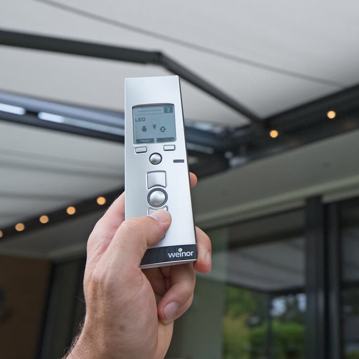 Eine Hand hält eine Fernbedienung vor einer Überdachung mit hellen Farben und modernen Linien.