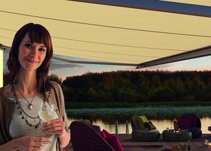Frau mit einem Getränk in der Hand, unter einer Markise, im Hintergrund ein Gewässer und sitzende Terrassensituation. 