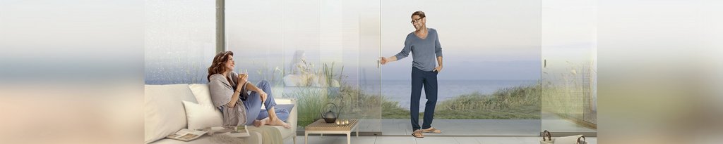 Zwei Personen in einer modernen Terrassensituation mit Glas-Schiebewänden und Ausblick auf das Meer. Zwei Personen in einer modernen Terrassensituation mit Glas-Schiebewänden und Ausblick auf das Meer.