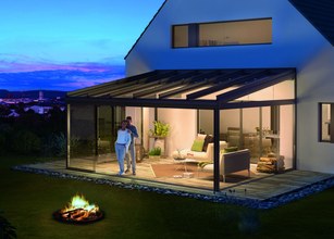 Markise über einer gemütlichen Terrassensituation mit Glas-Schiebewänden und einer Feuerstelle im Vordergrund. 